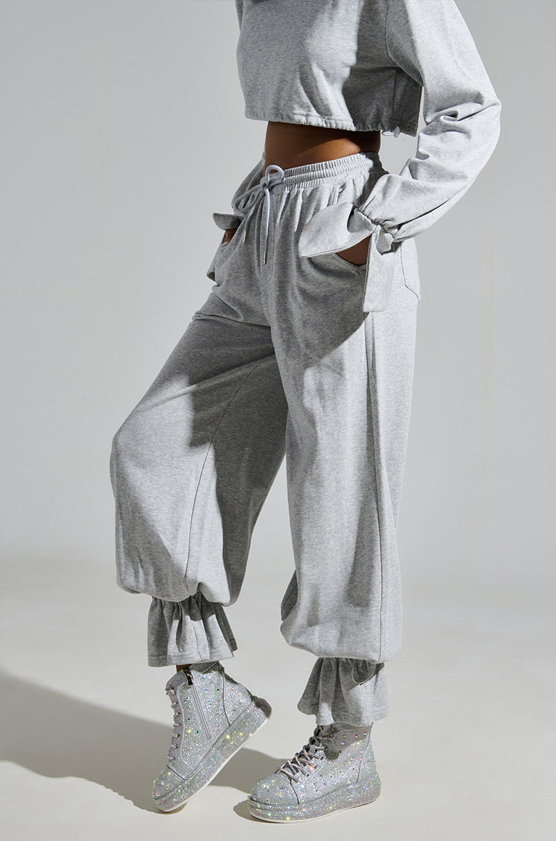 Wearing a gray sweat set with a cropped top and wide-leg pants with ankle ruching, they pair the outfit with AZALEA WANG OTIS Rhinestone High Top Sneakers in Silver. Hands in pockets create a casual, stylish pose.