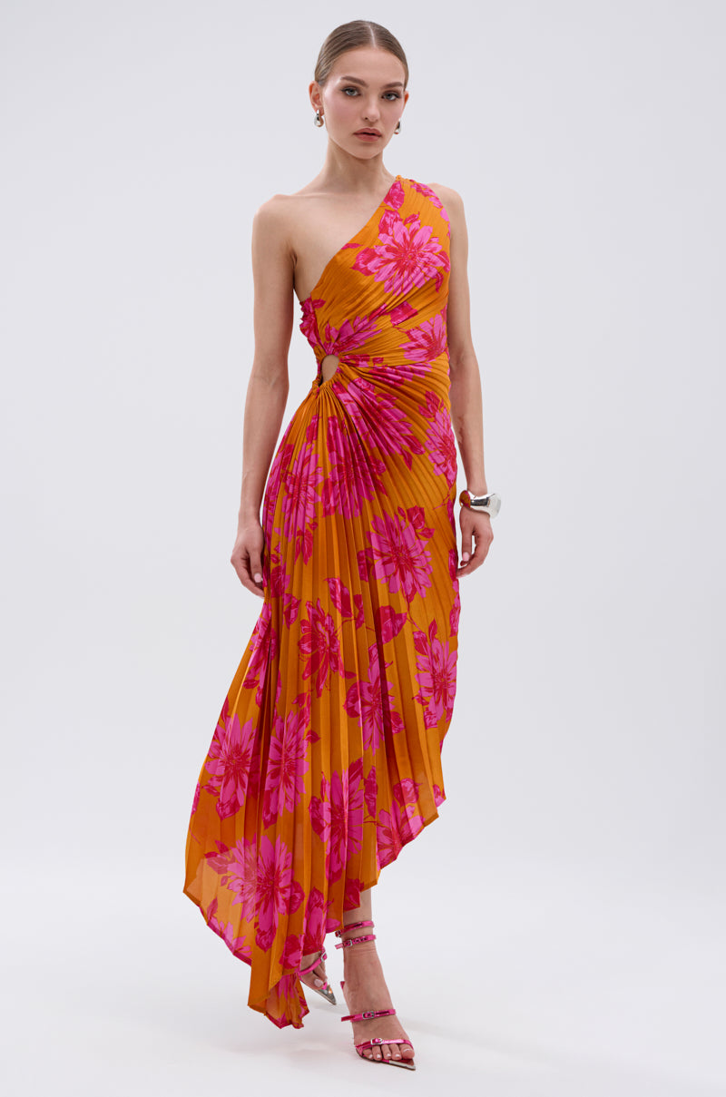 A woman wears the RADIANT ENERGY PRINTED MIDI DRESS, a bright orange asymmetrical midi with tropical floral print and pleats, styled with pink strappy heels and a white bracelet, standing against a plain white background.