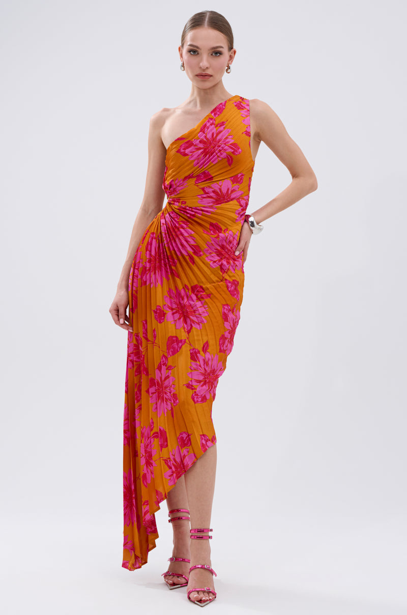 A woman in the RADIANT ENERGY PRINTED MIDI DRESS—a bright orange midi dress with a tropical floral print and asymmetrical design—pairs it with pink strappy heels and hoop earrings while posing against a white background.