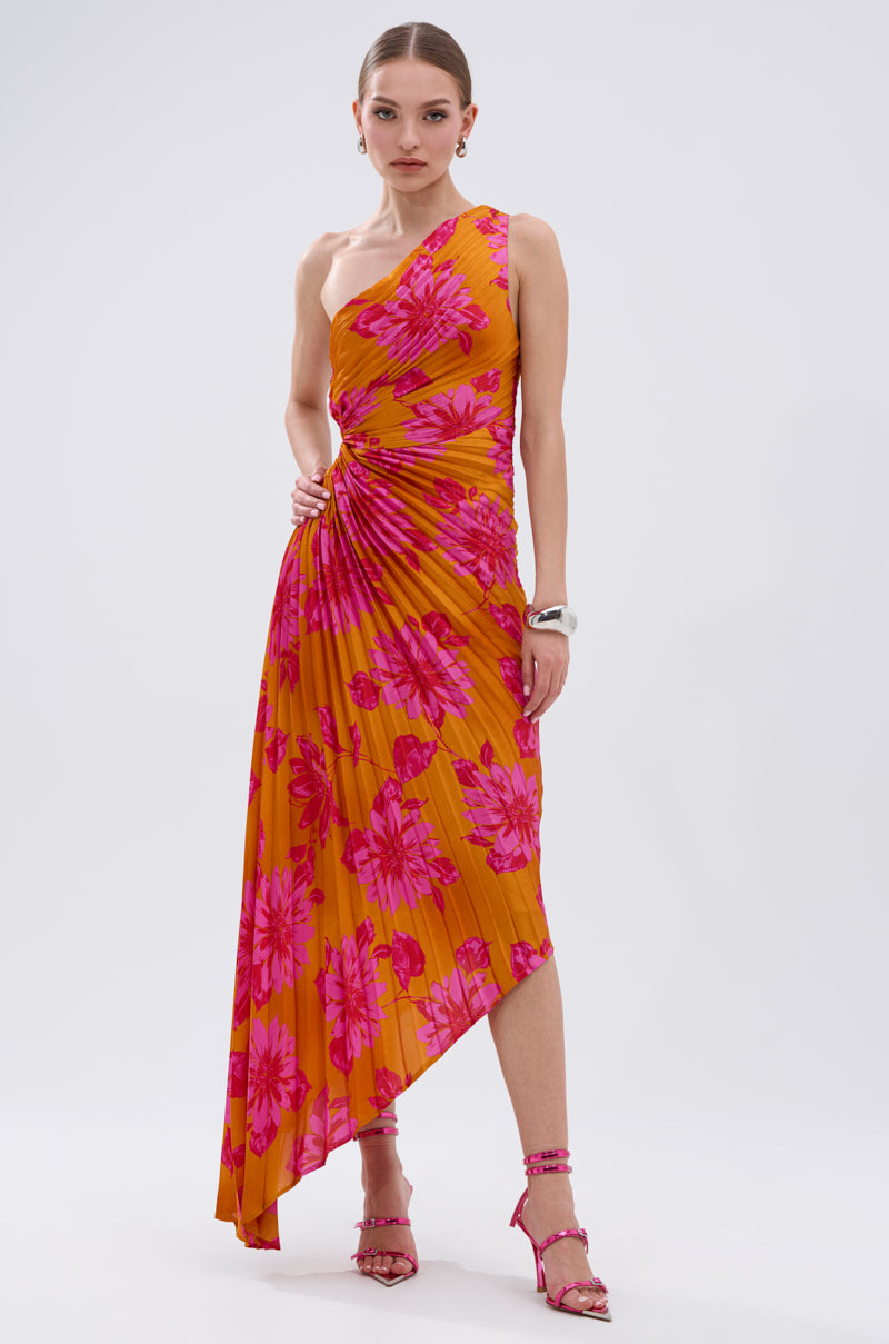 A woman wears the RADIANT ENERGY PRINTED MIDI DRESS—an asymmetrical midi with bold tropical floral print and pleats—paired with pink strappy heels and silver accessories, standing against a plain white background.
