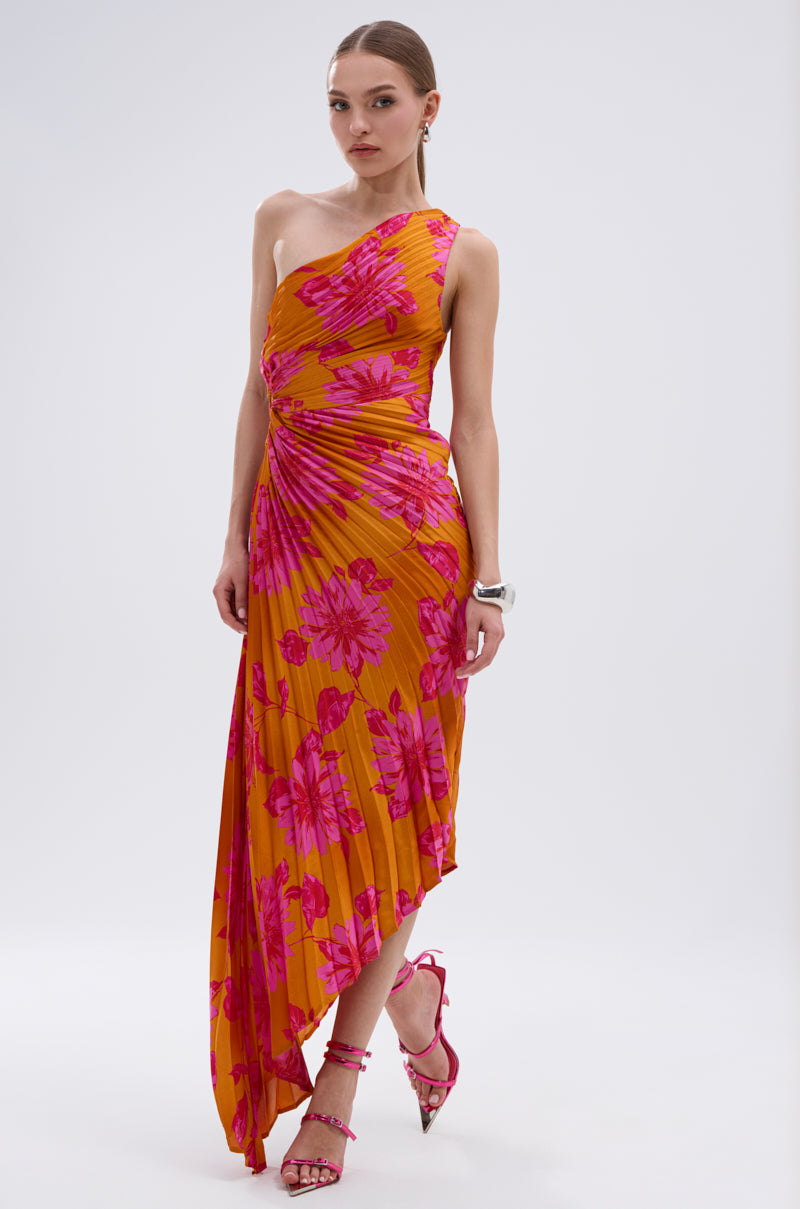 A woman wears the RADIANT ENERGY PRINTED MIDI DRESS, an orange asymmetrical midi with a tropical floral print, paired with pink strappy heels and a white bracelet as she stands against a plain white background.