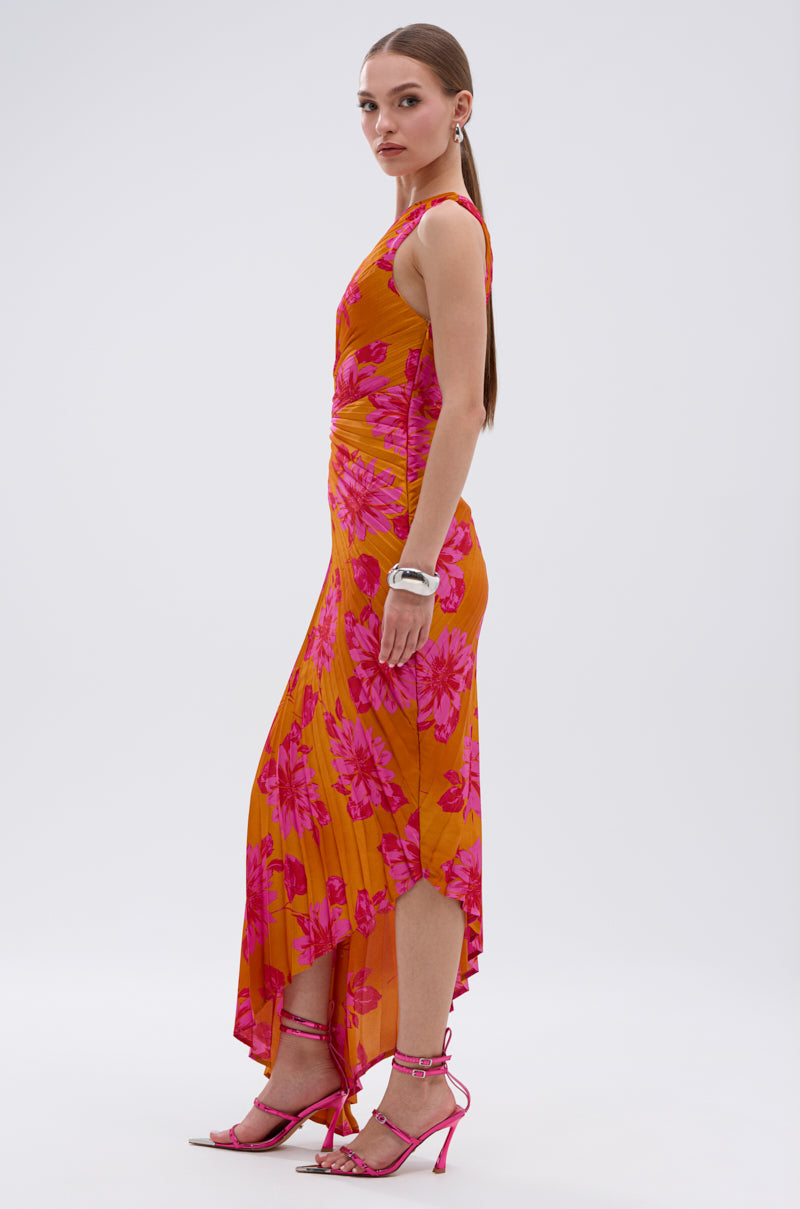 A woman stands sideways wearing the RADIANT ENERGY PRINTED MIDI DRESS, a sleeveless orange midi with vibrant tropical floral print, paired with pink heels and silver jewelry, against a plain light background.