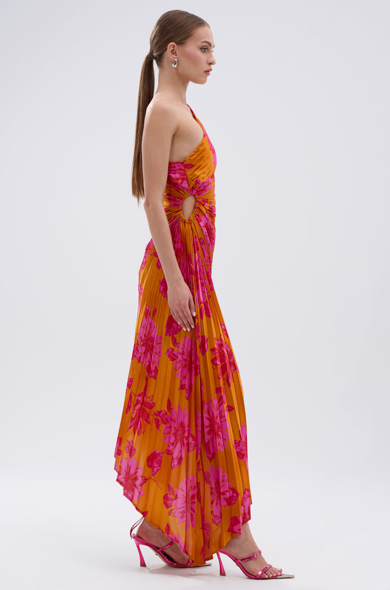 A woman with long brown hair in a low ponytail wears the RADIANT ENERGY PRINTED MIDI DRESS, featuring a vibrant tropical floral print and side cut-outs, paired with pink heeled sandals, standing in profile against a light background.