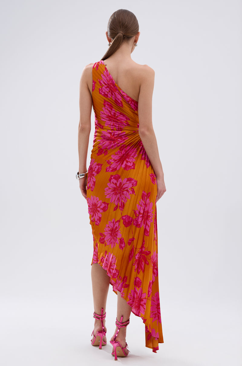 A woman with long hair in a low ponytail wears the RADIANT ENERGY PRINTED MIDI DRESS, a one-shoulder orange midi dress with a vibrant tropical floral print. She stands with her back to the camera, paired with pink high-heeled sandals.