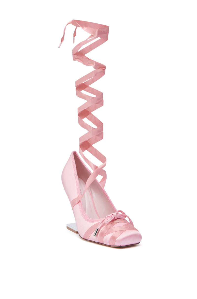 The AZALEA WANG PAVARTI PINK BALLET PUMP features a satin upper, inverted wedge heel, and crisscross satin ribbons in a zigzag pattern, evoking the look of a ballerina’s pointe shoe.