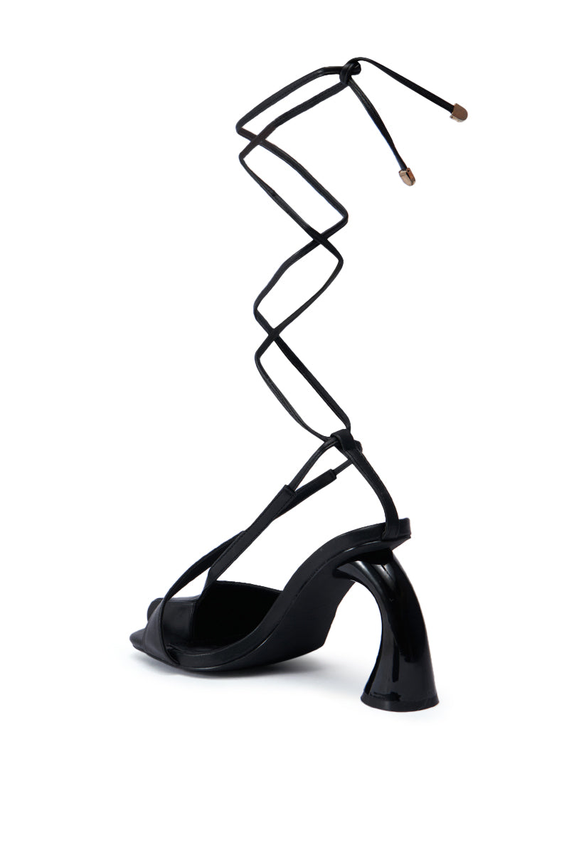 The PIRATE BLACK PUMP is showcased with a distinct, sculptural heel and a long, thin strap that elegantly wraps around the ankle. The strap is finished off with two small metal caps, adding to its modern and artistic design. This strappy sandal truly makes a statement.