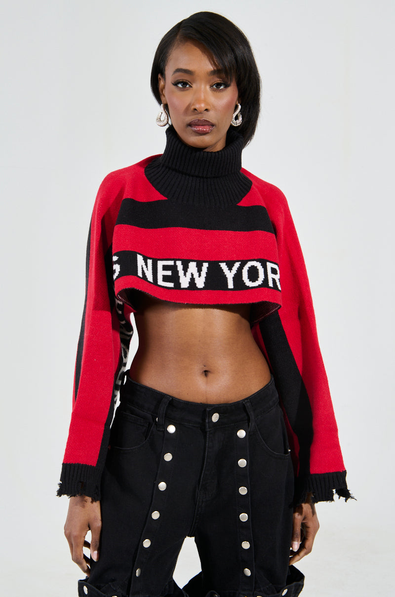 Someone poses in a chic NEW YORK ULTRA CROP SWEATER IN RED MULTI with an oversized turtleneck, showcasing a red and black colorblock design. They wear high-waisted black pants with multiple buttons, set against a plain white background.