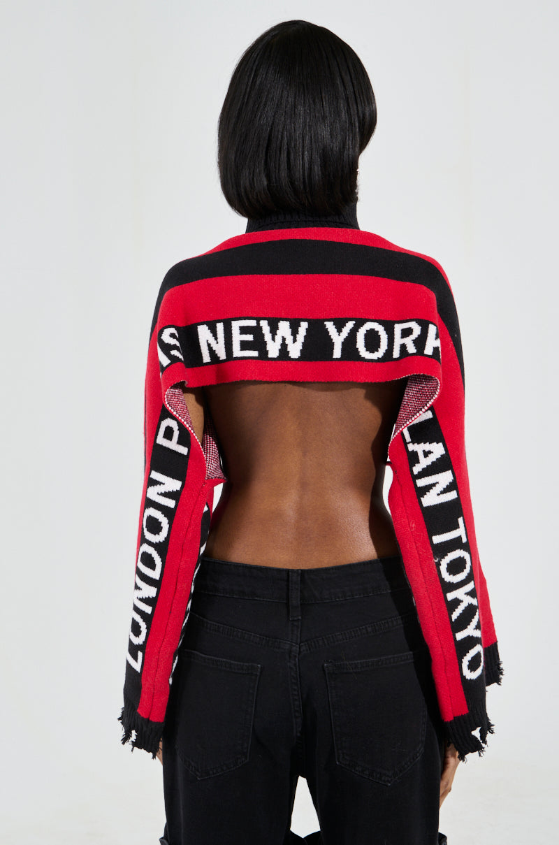 A person with shoulder-length black hair wears a striking New York Ultra Crop Sweater in Red Multi, featuring "NEW YORK" and "TOKYO" on a bold colorblock pattern, casually draped to reveal their back. Paired with sleek black pants, they effortlessly exude urban chic.