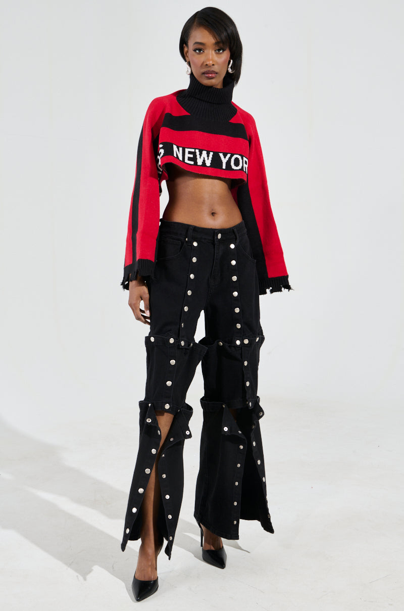 A model poses against a plain background, wearing a vibrant New York Ultra Crop Sweater in Red Multi and unique black pants with button accents and flared openings, completed with black high heels.