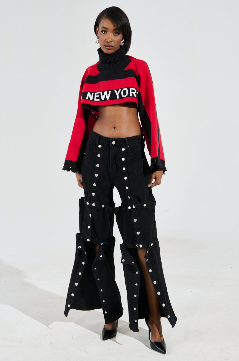 A person wears the New York Ultra Crop Sweater in Red Multi, featuring a chic colorblock pattern. They pair it with black wide-leg pants with button details. Standing confidently against a white background, their shoulder-length hair and hand stylishly on their hip complete the look.
