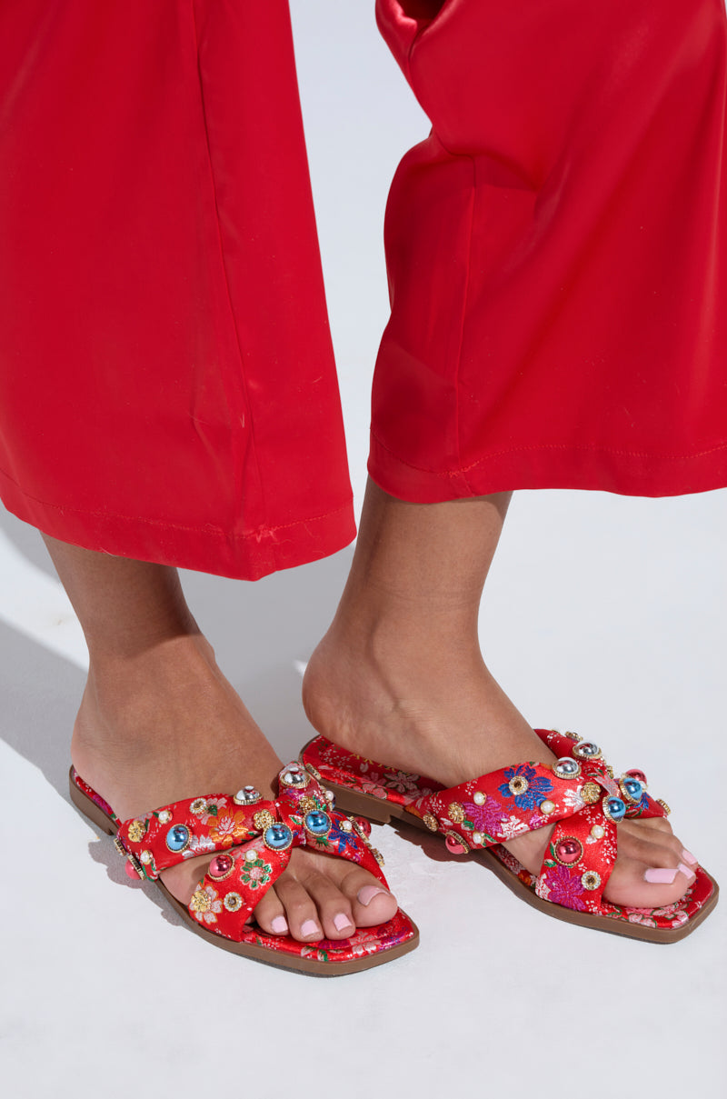 Someone in vibrant red pants complemented by the AKIRA RAMSEY RED FLAT SANDALS, featuring floral embellishments and cushioned insoles. The well-lit setting accentuates the sandals' bright colors and detailed patterns.