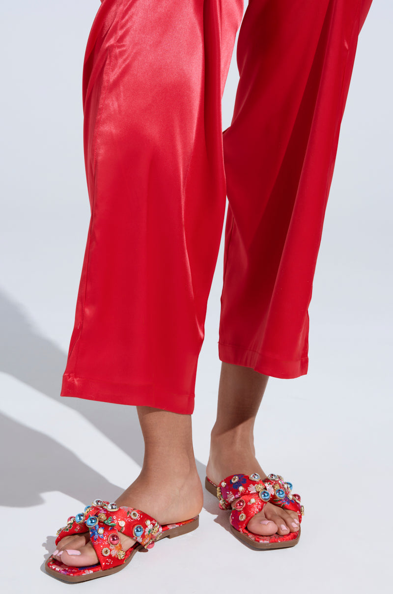 A person wearing red satin pants and the AKIRA RAMSEY RED FLAT SANDAL, adorned with floral embellishments and colorful gems, complements the sheen of the pants against a neutral, minimalistic background.