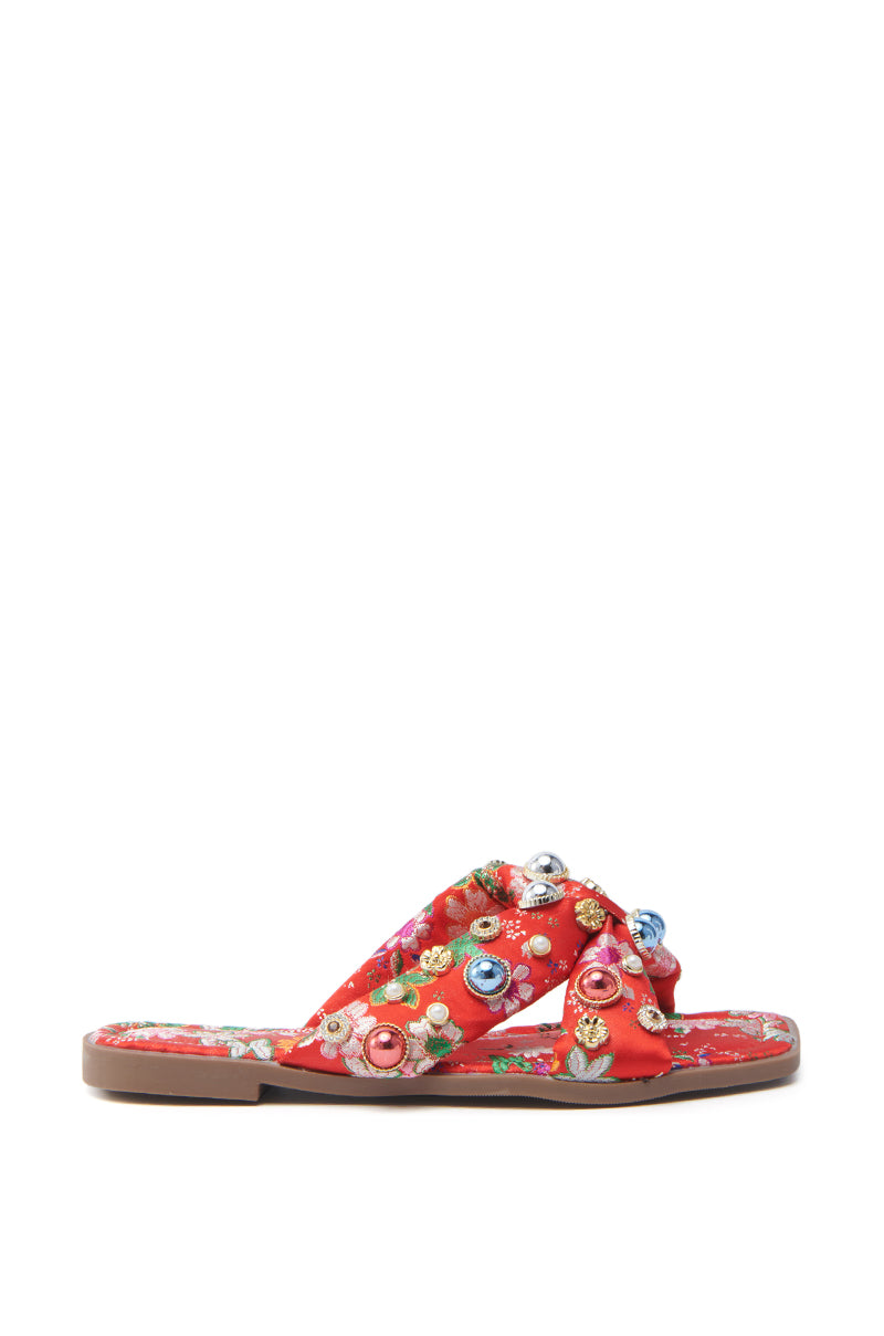 The AKIRA RAMSEY RED FLAT SANDAL features red satin with a flat sole, woven crisscross straps adorned with colorful floral embroidery, embellishments, and eyelet accents. It includes a cushioned insole for added comfort.