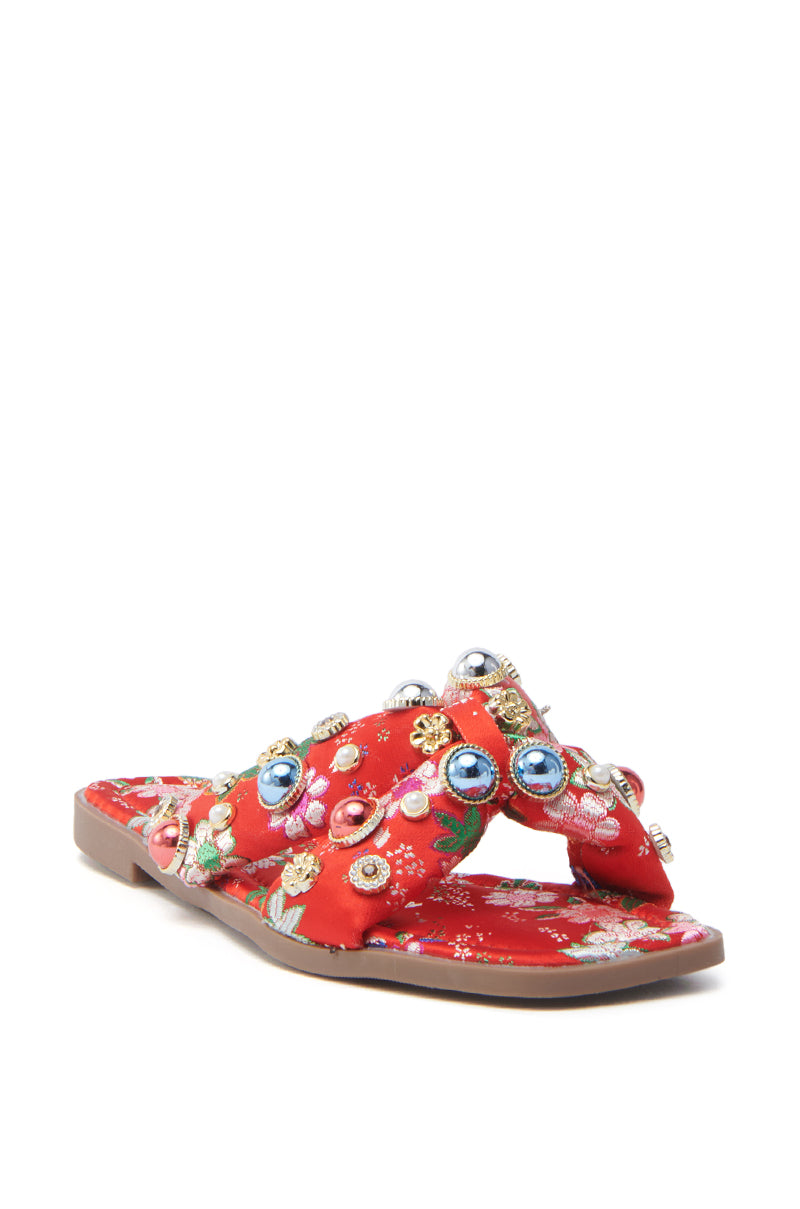 The AKIRA RAMSEY RED FLAT SANDAL is a red satin sandal with crisscross straps, floral embellishments, and colorful beads in blue, pink, and gold on a flat brown sole. Its ornate design offers a festive look with a cushioned insole for comfort.
