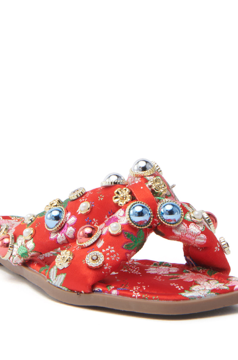 The AKIRA RAMSEY RED FLAT SANDAL is a vibrant red satin sandal featuring floral embellishments, crisscross straps adorned with beads and metallic studs. A cushioned insole ensures comfort while its decorative pattern provides an eye-catching and elegant design.