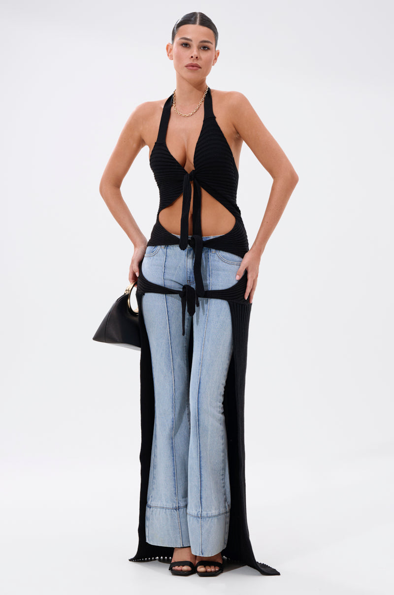 A woman poses against a plain background wearing the TIED TO YOU TIE-FRONT SWEATER with front cutouts, light blue wide-leg jeans, a long black overlay, black sandals, and carrying a black handbag.