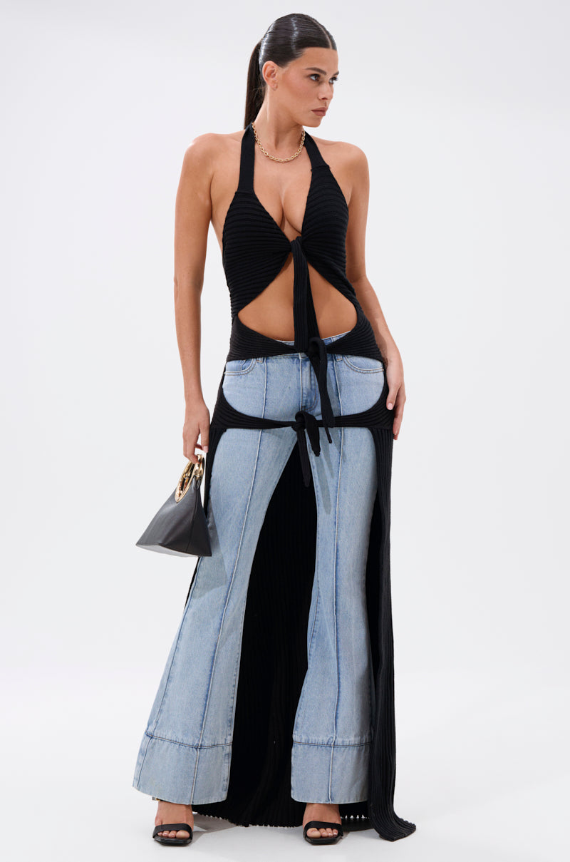 A woman poses in the TIED TO YOU TIE-FRONT SWEATER, styled with flared two-tone pants and light denim panels over a black skirt. She accessorizes with a small black handbag and black high-heeled sandals for a sleek longline look.