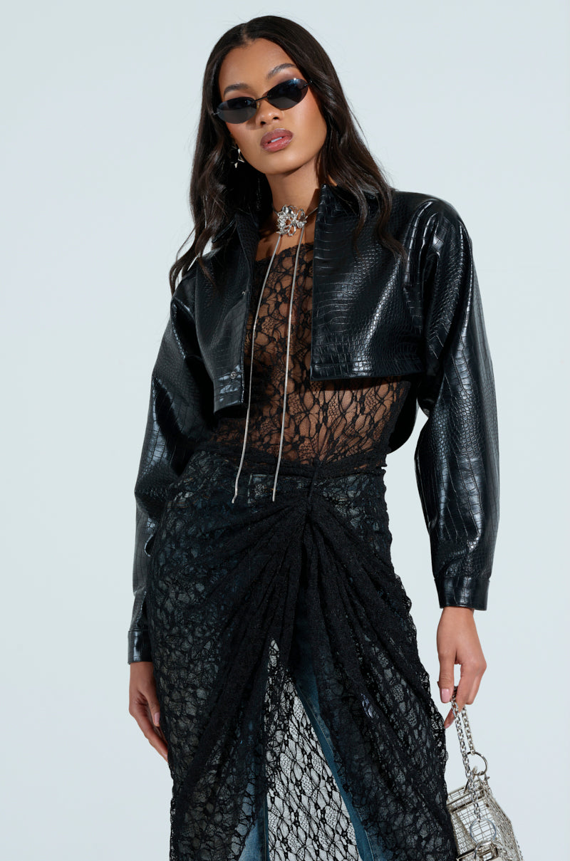 A woman wearing dark sunglasses poses confidently in a stylish outfit. Her black leather jacket complements a transparent lace top and long skirt over pants, paired with bold jewelry. She carries a textured handbag set against a plain light background, accentuating her NATALIA CROCODILE PU BUTTON DOWN SHIRT.