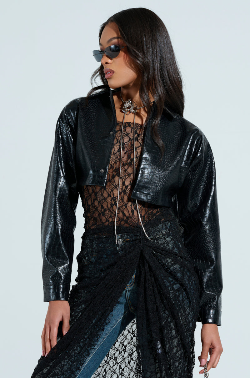 A person models a chic, edgy look in black sunglasses and the NATALIA CROCODILE PU BUTTON DOWN SHIRT, paired with a sheer lace top and a blue denim skirt with black lace overlay. Their long dark hair falls loosely as they pose slightly to the side, highlighting a fashionable silhouette.