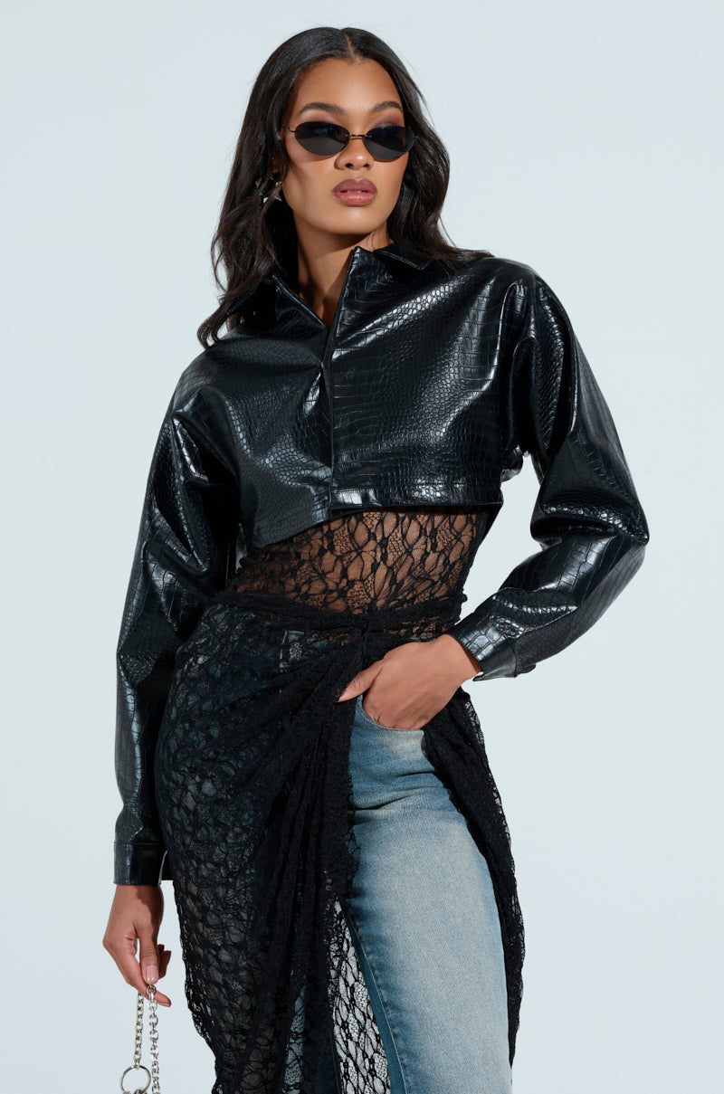 A woman poses confidently in a stylish outfit featuring the NATALIA CROCODILE PU BUTTON DOWN SHIRT with a black netted overlay, paired with blue jeans. She wears dark sunglasses and has long, wavy hair.