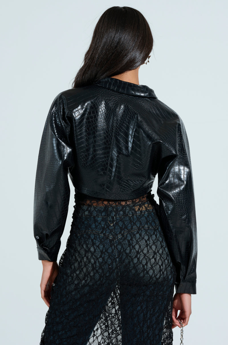 A person is seen from behind wearing the shiny black Natalia Crocodile PU Button Down Shirt and a long, sheer lace skirt with a shorter lining underneath. The cropped silhouette of the shirt adds an edgy touch against the plain, light-colored background.