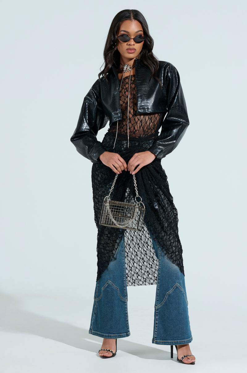 A person poses confidently in a stylish outfit featuring a cropped silhouette with sunglasses, wearing the NATALIA CROCODILE PU BUTTON DOWN SHIRT under a black faux leather jacket, flared denim jeans, and a sheer lace top peeking through. They hold a metallic purse and wear strappy heels.