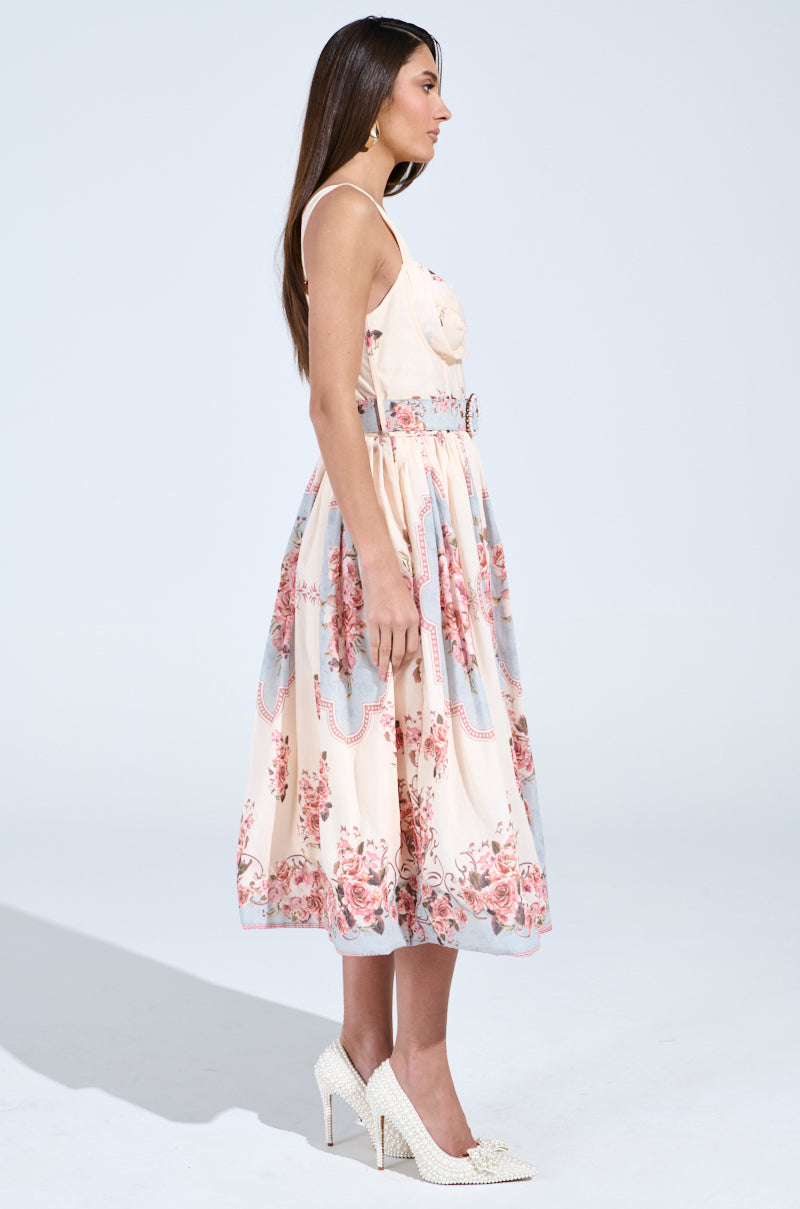 A woman in profile models the sleeveless HANNA FLORAL MIDI DRESS, featuring a vintage-inspired floral pattern and a mid-length skirt. Her long hair cascades gracefully against a light background, complemented by studded heels and a belt reflecting the dress's charm.