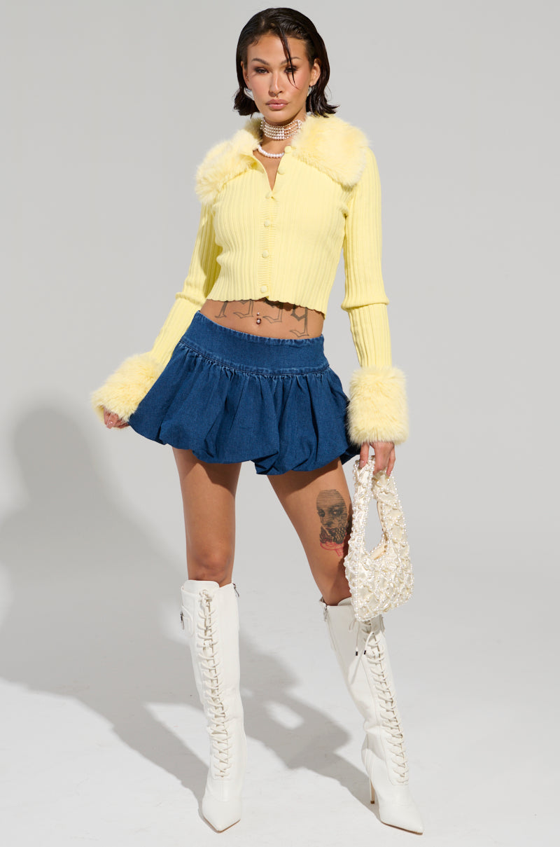 A person poses confidently in a yellow fur-trimmed cardigan and the PRETTY WOMAN DENIM BUBBLE SKIRT. They're holding a lace handbag and wearing white knee-high boots. Their outfit is accented with various jewelry, and they stand against a plain light background.
