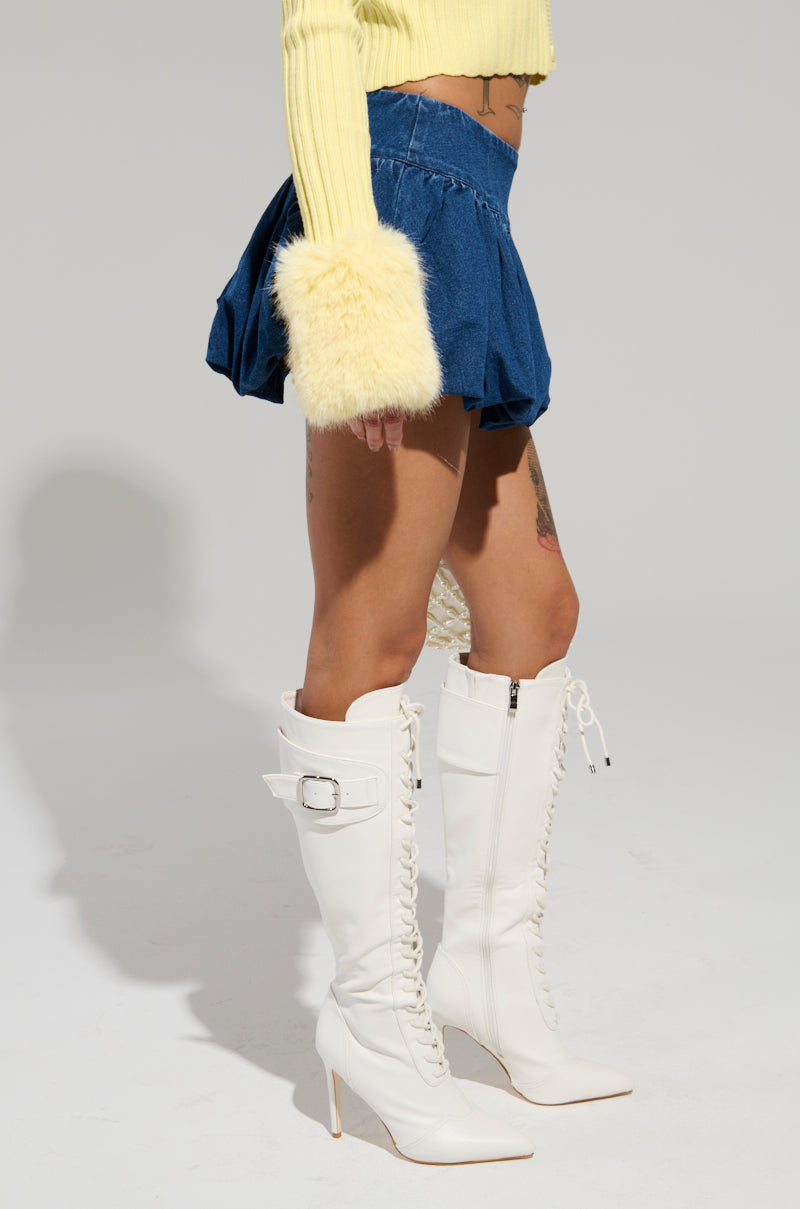 A person dons a yellow sweater with fuzzy cuffs, paired elegantly with the PRETTY WOMAN DENIM BUBBLE SKIRT, a high-rise mini-length denim piece. Completing the chic ensemble are white knee-high lace-up boots with high heels, all captured in a stylish pose from mid-torso.