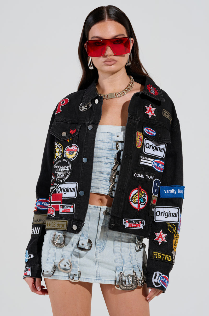 A woman models the "PATCH IT UP DENIM JACKET" featuring letterman patches, paired with a light blue cropped top and a mini skirt accented with metallic rings. She completes her look with long dark hair, red sunglasses, and a chunky chain necklace, set against a plain background.