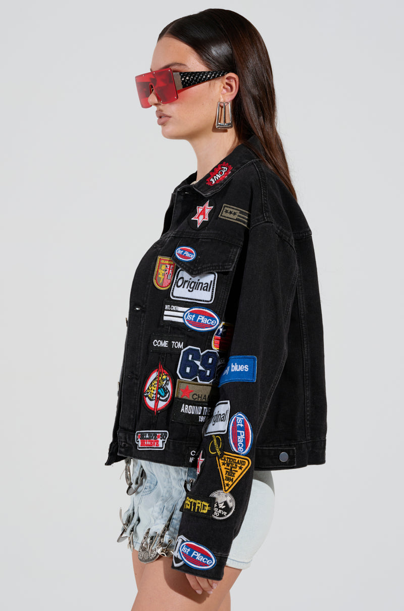 Wearing the PATCH IT UP DENIM JACKET adorned with vibrant letterman patches and symbols, a person sporting oversized red sunglasses, large earrings, and light-colored shorts that create a chic cropped silhouette stands sideways. Their long hair cascades over the jacket.
