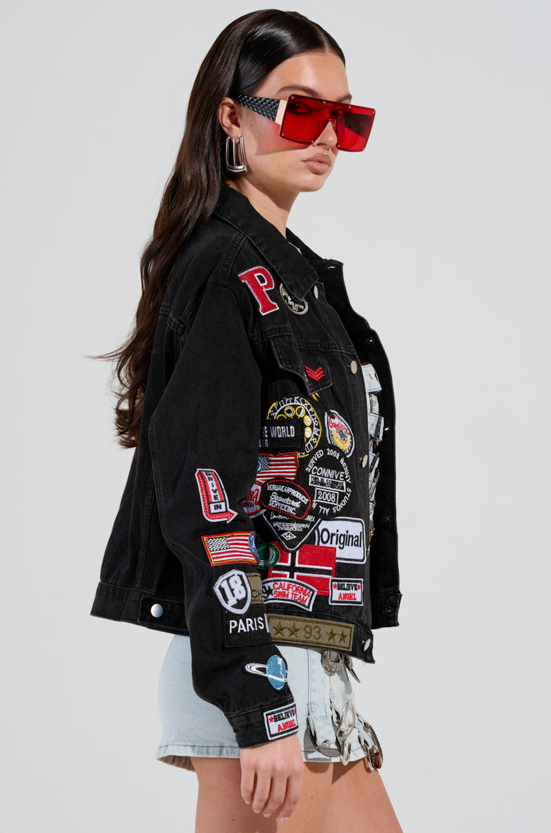 A person wears the PATCH IT UP DENIM JACKET, embellished with vibrant letterman patches. They accessorize with large red sunglasses and light blue shorts, standing against a plain background. The jacket boasts a distinctive mix of logos, flags, and text for a bold look.