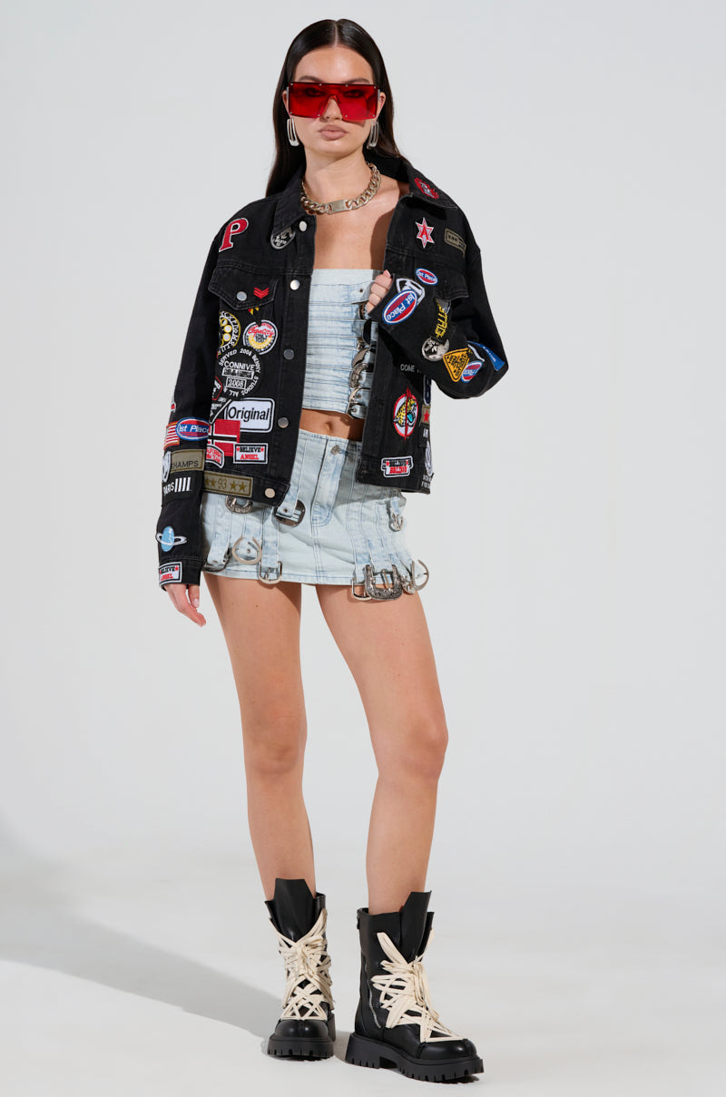 A woman stands confidently in a denim skirt and tube top, layered with the PATCH IT UP DENIM JACKET, adorned with colorful letterman patches. She accessorizes with red sunglasses, a chunky chain necklace, and striking black boots with cream accents.