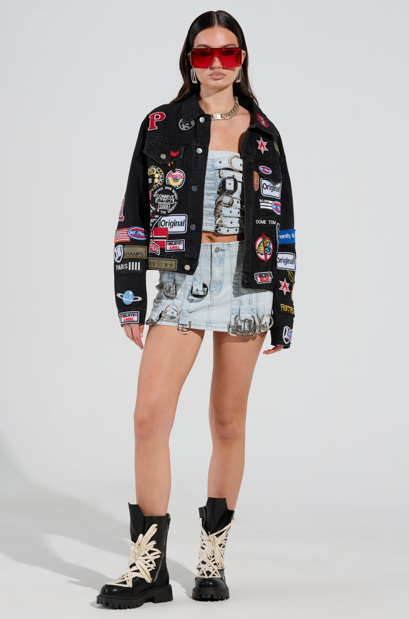 Dressed in the PATCH IT UP DENIM JACKET featuring letterman patches, an individual pairs it with a light mini skirt and crop top. They accessorize with red sunglasses, a choker necklace, and black lace-up boots with thick soles to create a bold and edgy appearance.