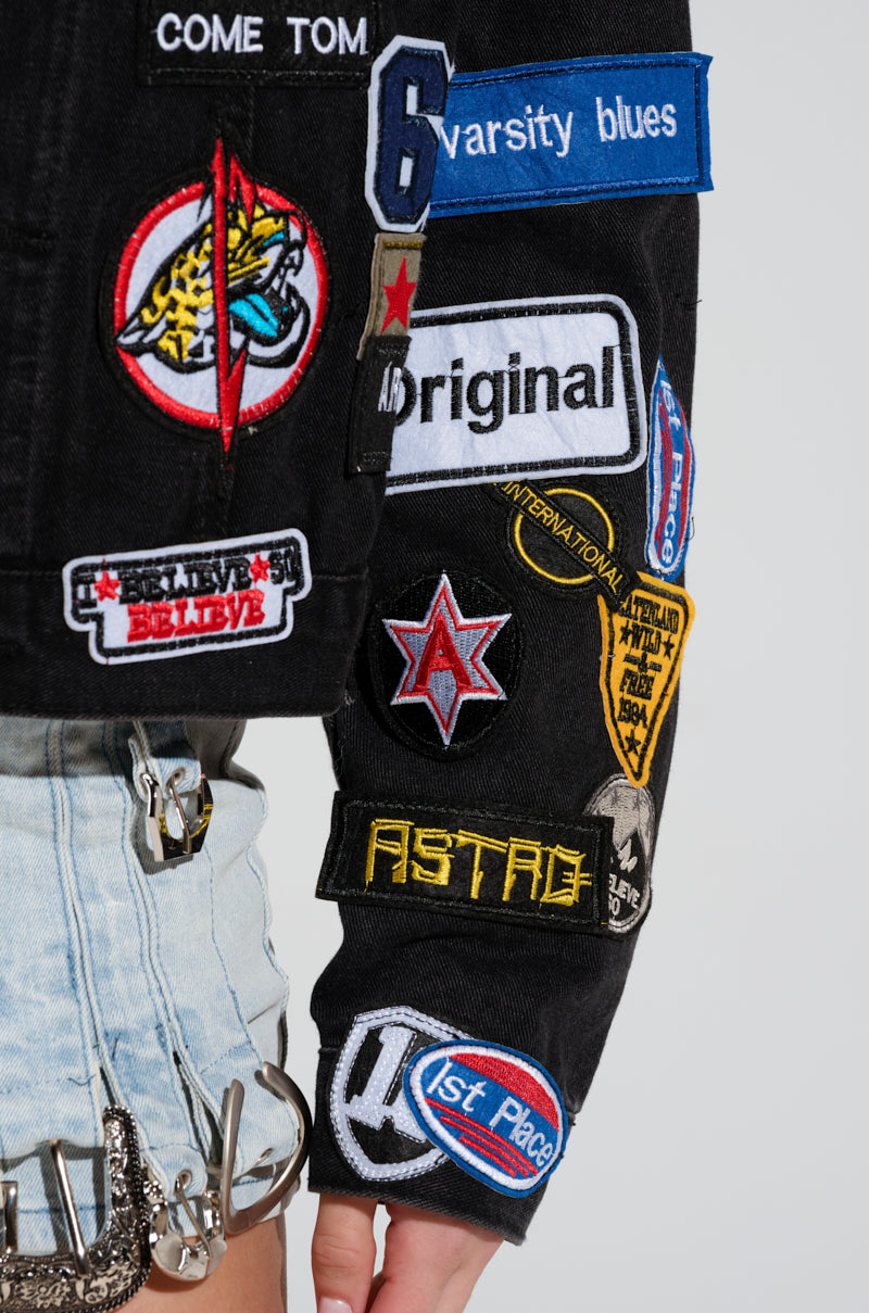 A person wearing the PATCH IT UP DENIM JACKET, a black cropped silhouette with vibrant letterman patches featuring phrases such as "International," "Varsity Blues," and "Astro." It's paired with light denim shorts decorated with metal chain details.