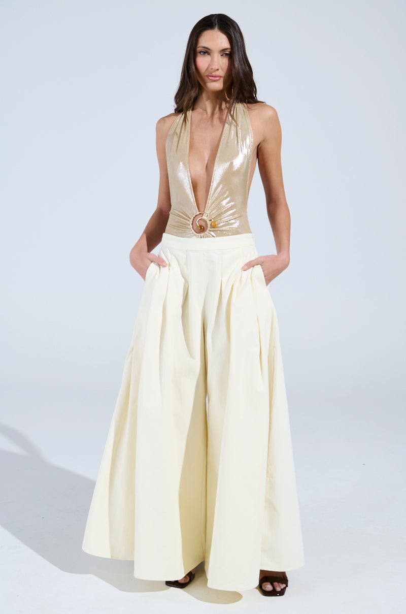 In a studio, a woman confidently wears a shiny gold halter top with a deep V-neck and circular buckle. She pairs this with TAY TAY TROUSERS, showcasing an ultra-wide-leg silhouette and mid-rise fit. Her hands rest in her pockets, completed by sleek black heels.