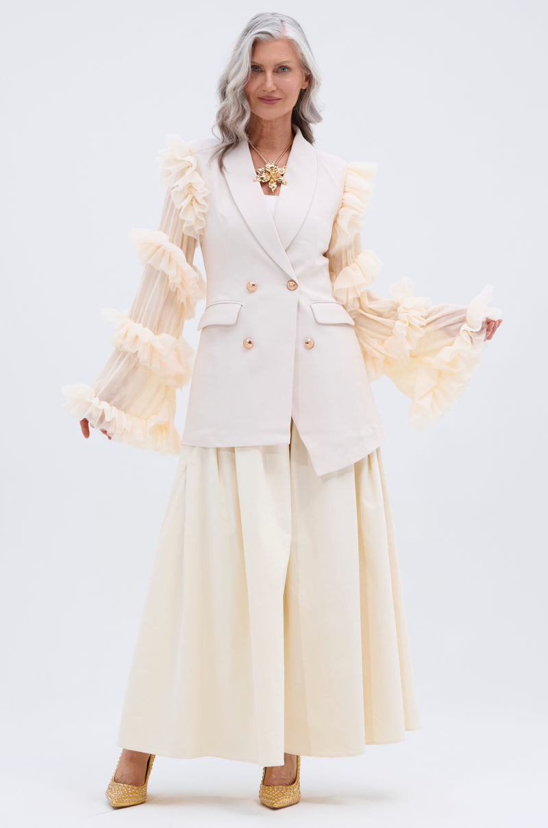 A woman with long gray hair stands confidently against a white background wearing the TAY TAY TROUSER, a cream blazer with sheer, ruffled sleeves, a flowing pleated ivory skirt, gold necklace, and heels.