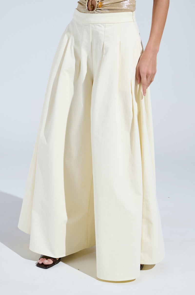 The individual wears high-waisted, wide-leg cream pants with pleats from the AKIRA Label's TAY TAY TROUSER collection. They pose confidently in a gold sequined top and black open-toe shoes, with one hand on their hip.
