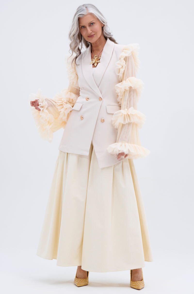 A woman with long gray hair stands confidently in yellow heels against a white background, wearing a cream double-breasted vest with sheer ruffled sleeves and the pale yellow TAY TAY TROUSER in a wide-leg silhouette.