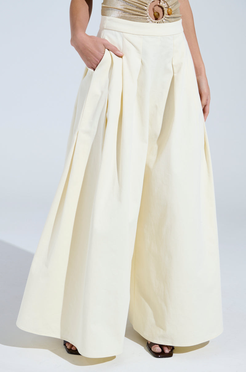 A person in mid-rise, wide-leg TAY TAY TROUSER in cream pleats stands with a hand in one pocket. They wear a beige belt and top with gold ring detail. The floor-length trousers give just a peek of the footwear underneath.