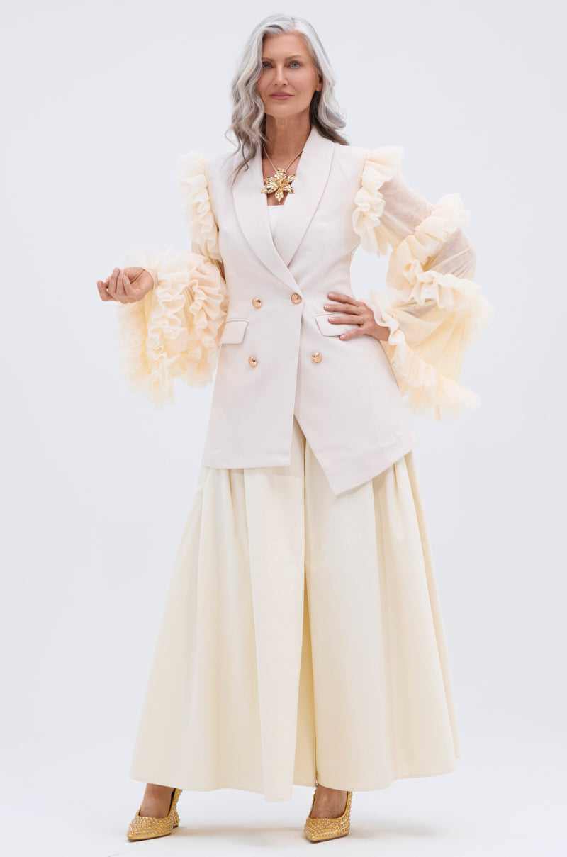 A stylish older woman with long gray hair confidently models the TAY TAY TROUSER—a flowy, cream wide-leg pant—paired with a cream vest featuring ruffled sheer sleeves, gold shoes, and a statement necklace against a white background.
