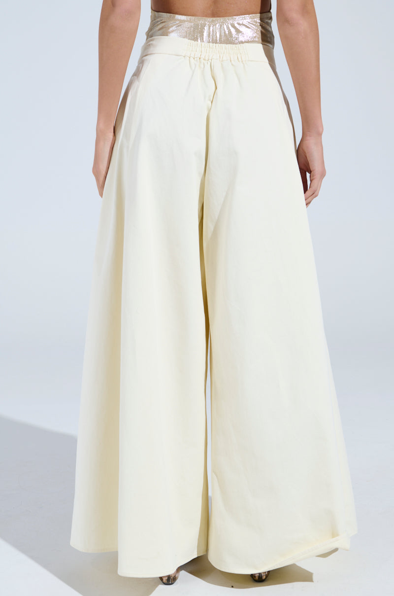 The individual is wearing high-waisted, cream AKIRA Label TAY TAY TROUSERs with an ultra-wide-leg and shimmering gold waistband, set against a plain, light background as they face away from the camera.