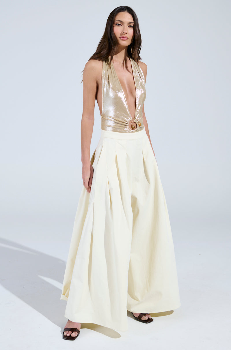 A woman exudes confidence in a shiny gold halter top and TAY TAY TROUSER with an ultra-wide-leg design. Her long brown hair flows freely as she gazes at the camera against a light background, finishing her look with dark open-toed shoes.