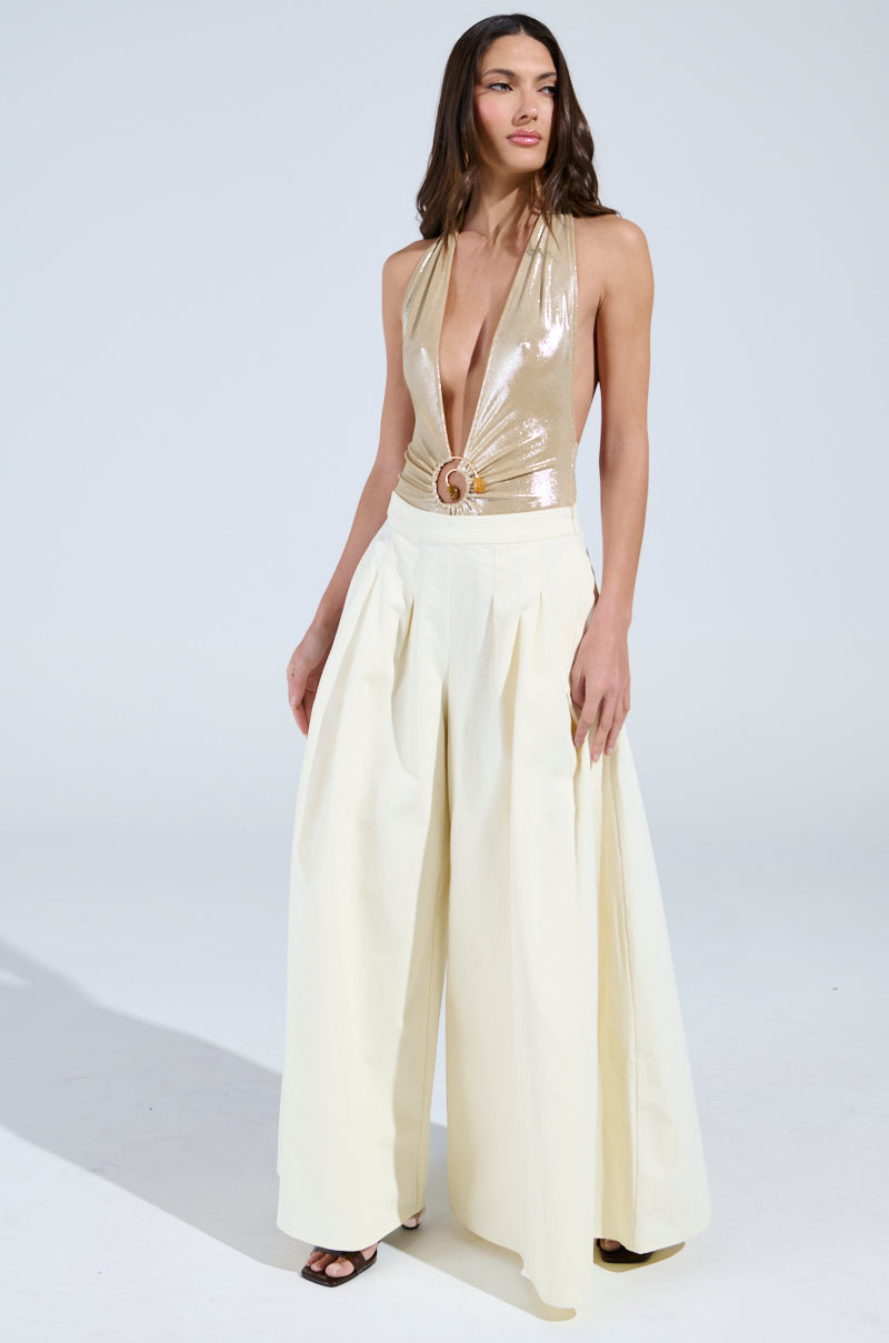 A woman stands confidently against a plain background, wearing a shiny gold sleeveless top with a deep V-neckline and a circular embellishment at the waist. She pairs it effortlessly with the TAY TAY TROUSER, high-waisted and wide-leg in cream, along with black open-toe shoes.