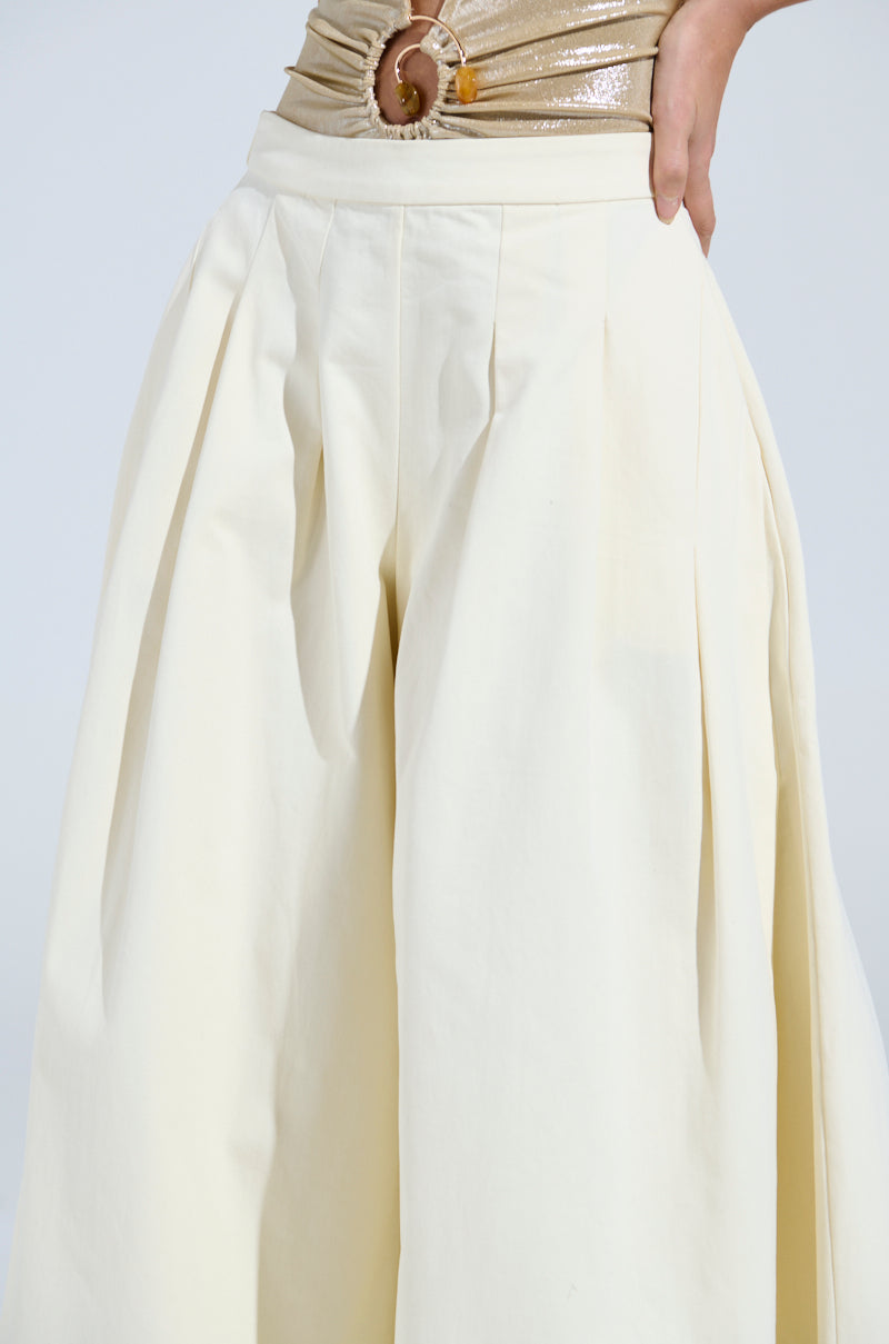 Someone is wearing the TAY TAY TROUSER, which are high-waisted, wide-leg cream pleated trousers, paired with a shiny gold top featuring a circular clasp at the waist against a plain neutral background.