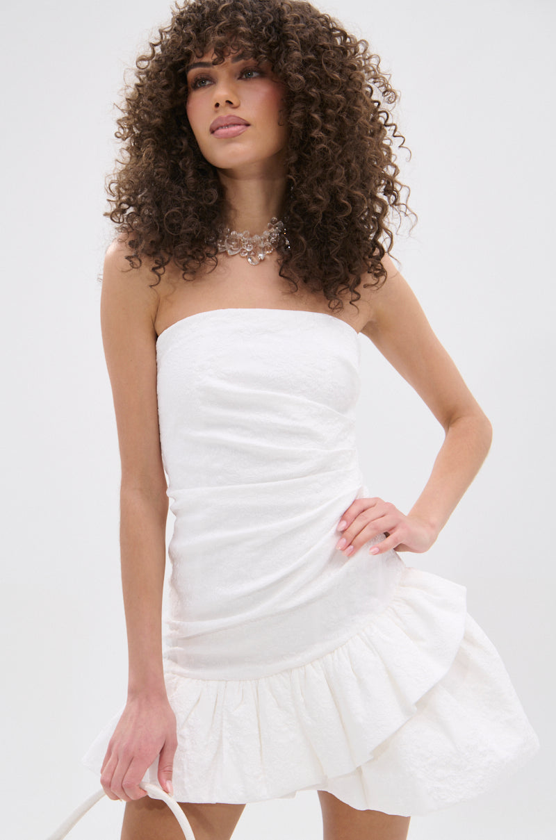 A woman with curly hair wears the GARDEN PARTY MINI DRESS IN WHITE, featuring a strapless design and ruffled hem, perfect for a garden party. She pairs it with a statement necklace against a plain light background.
