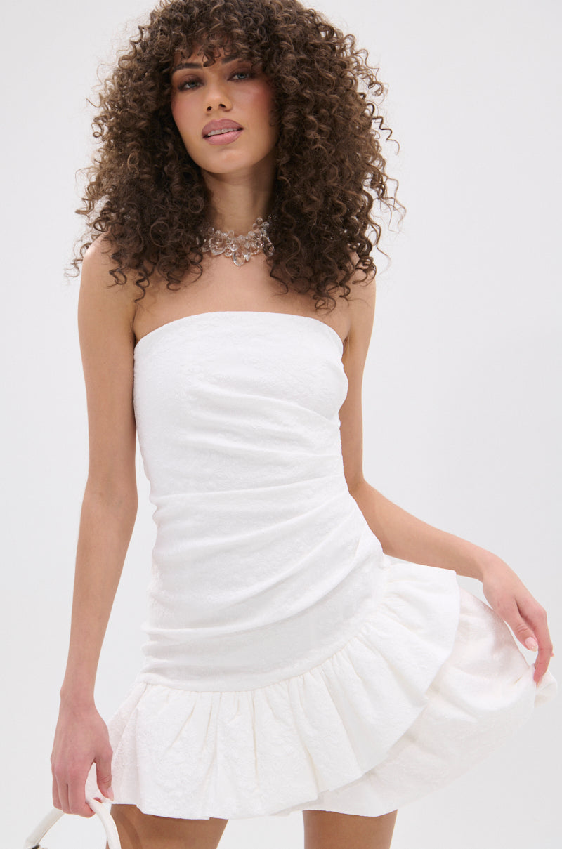 A woman with curly hair poses against a light background in the GARDEN PARTY MINI DRESS IN WHITE, a strapless mini dress with a ruffled hem. She pairs it with a beaded necklace and holds the skirt, ready for a chic garden party look.