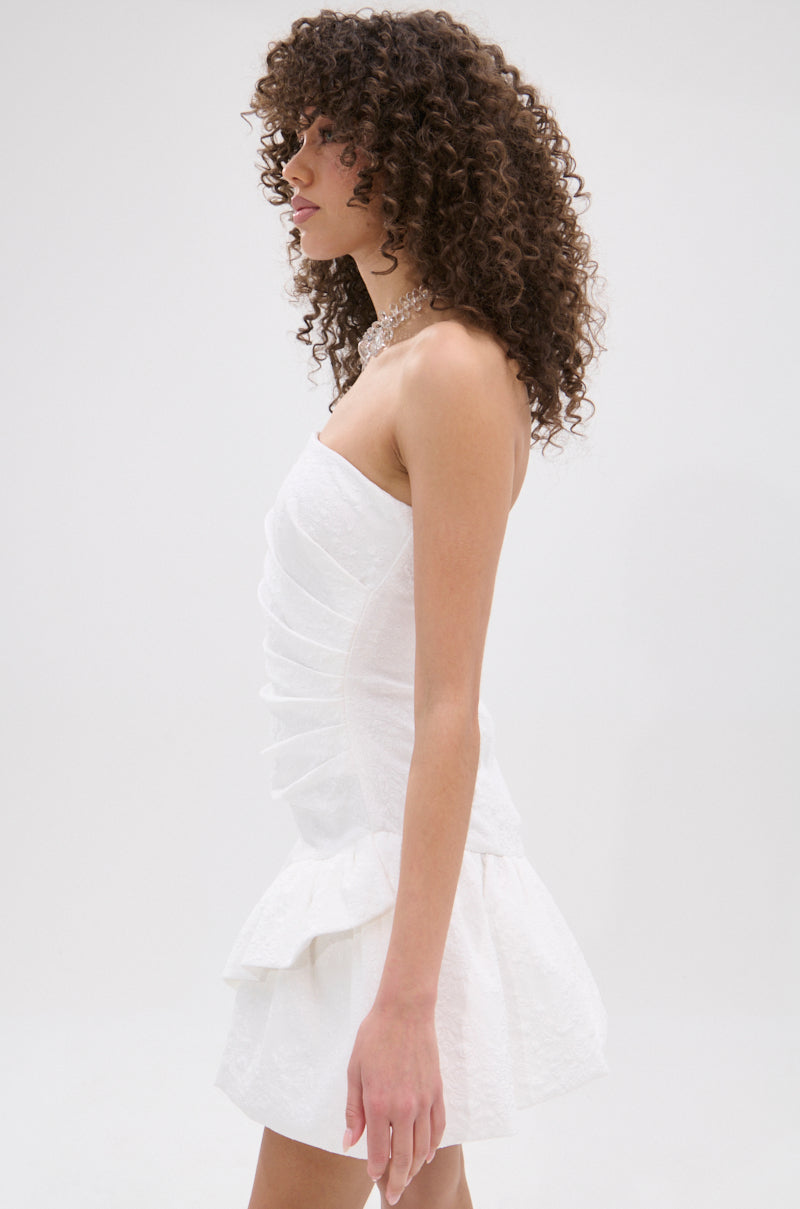 A woman with curly hair models the GARDEN PARTY MINI DRESS IN WHITE—a strapless, textured mini dress with a fitted bodice and ruffled skirt—posing in profile against a plain light background, ideal for chic garden party style.