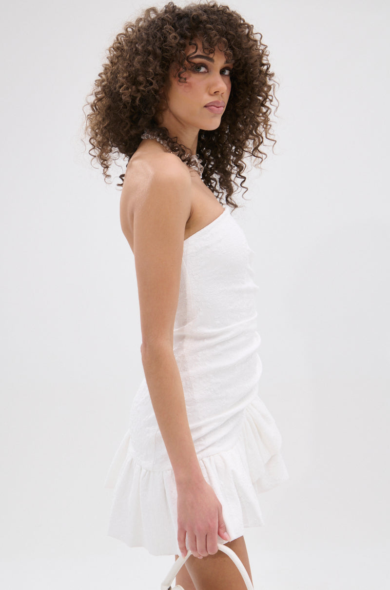 A woman with curly hair models the GARDEN PARTY MINI DRESS IN WHITE, featuring a strapless floral print and ruffled hem, standing sideways against a white background while holding a white strap and glancing toward the camera.