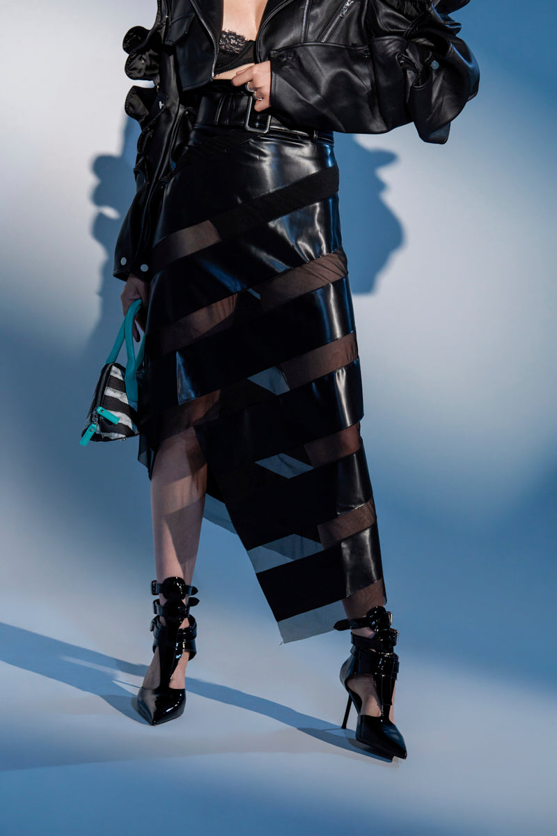 A person wears a black leather jacket over a lace top, the SOMETHING DIFFERENT MESH MIDI SKIRT WITH FAUX LEATHER, pointed buckled high heels, and holds a patterned purse.